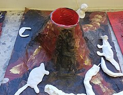Ein Vulkan aus Pappmaché. Innenleben ist eine Blechdose. Er ist mit Acrylfarbe angemalt. Aus Ton gefertigte Tiere, Vulkanbewohner, sitzen auf dem Boden.