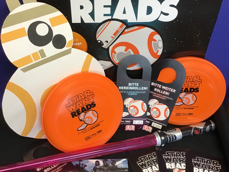Star-Wars-Reads-Day-Plakat mit BB8 daneben, Star-Wars-Give-Aways zB Frisbees, Lesezeichen, Postkarten, ein Lichtschwert.