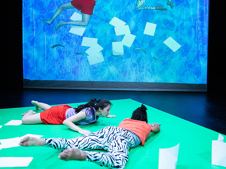 Zwei Schauspielerinnen liegen auf einer grünen Matte. Im Hintergrund ist eine Leinwand, auf der sie in ein Bild hineinprojiziert wurden