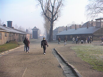 Blick in die heutige Gedenkstätte Auschwitz-Birkenau. Das ehemalige deutsche Konzentrationslager. Menschen bewegen sich über das Gelände im Hintergrund eine Baracke und ein Wachturm.