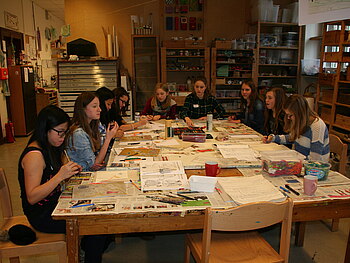 9 Mädchen im Teenager-Alter sitzen im Rahmen einer individuellen kreativen Geburtstagsfeier an einem Werktisch und gestalten - jede für sich - eine Mappe mit Modezeichnungen.
