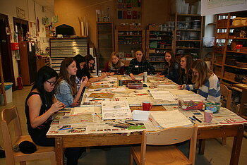9 Mädchen im Teenager-Alter sitzen im Rahmen einer individuellen kreativen Geburtstagsfeier an einem Werktisch und gestalten - jede für sich - eine Mappe mit Modezeichnungen.