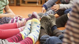 Füße von Kindergartenkinder, die im Kreis sitzen und die Füße aneinander halten. Manche Füße haben Stoppersocken über, andere Crogs