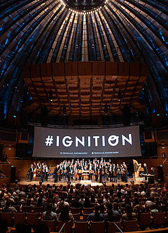 Stehendes Orchester auf Bühne unter Leinwand, worauf "Ignition" steht