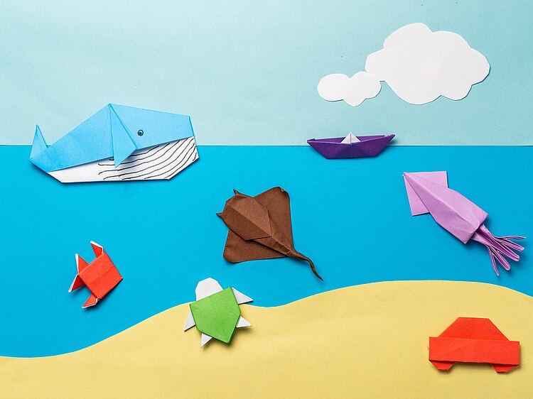 Eine Papiercollage: Strand, Meer, Himmel und weiße Wolken. Darin aus Papier gefaltet ein Wal, eine Schildkröte, ein Tintenfisch, ein Rochen, ein Fisch, ein Boot und am Strand ein kleines Auto
