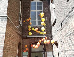 Ein hoher, schmaler Hauseingang mit einer hohen Tür und hohen Fenstern. In dem Eingang ist eine Kugelinstallation aus gelben und orangefarbenen Kugeln zu sehen. Die ca. 20 Kugeln haben die Größe von Luftballons.