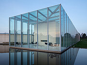 Außenansicht der Langen Foundation, ein postmoderner Bau mit Betonelementen und Glasflächen, sowie einer Wasserfläche in der Front.
