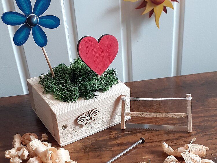 Ein kleiner Holzkasten, der liebevoll mit einem roten Holzherz, einer blauen Blume, etwas Moos und einem Holzschmetterling dekoriert ist.