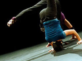 Ein Tänzer steht auf seinem Kopf und händen. Dahinter Gliedmaßen eines weiteren Tänzers in der gleichen Bewegung.