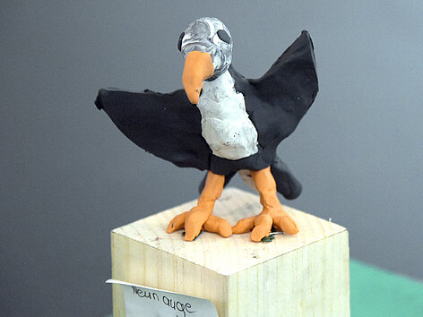Ein schwarz-weißer Vogel mit orangem Schnabel und orangen Beinen steht auf einem quadratischen hellen Holzsockel. Der Vogel ist aus Knete geformt.