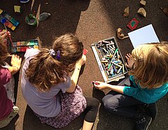 Drei Kinder sitzen draußen auf dem Boden und malen mit Wasserfarbe und Ölkreide Holzstücke an.