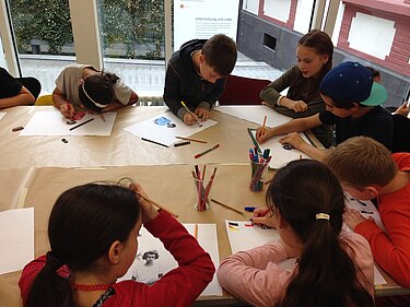 Im Anschluß an die Kinderführung gestalten sich Kinder Erinnerungsbilder. Die Kinder sitzen im Studio um einen abgerundeten Tisch und malen, basteln, zeichnen.