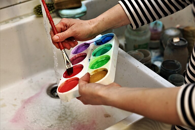 Hände die einen Pinsel im Waschbecken eines Künstlerateliers unter fliexend Wasser reinigen. 
