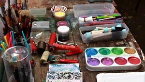 Ein Tisch mit verschiedenen künstlerischen Materialien wie Pinsel, Farbkasten und Walzrolle