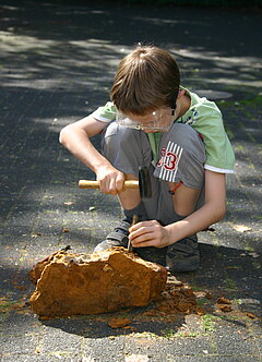 Grundschüler mit Schutzbrille auf kniet vor einem Gesteinsbrocken und bearbeitet ihn mit Hammer und Meißel.