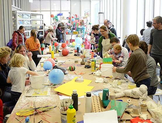 Workshop: Viele Kinder und Erwachsene an einem großen Tisch arbeiten gemeinsam an Skulpturen. Auf dem Tisch: Luftballons, dünne Holzstäbe, Kleber, farbiges Papier.