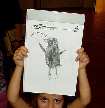 Ein Mächen im Grundschulalter hält ihre Zeichnung eines Pinguins in die Luft.