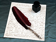Eine rote Schreibfeder liegt auf einem handgeschriebenen Brief. Oben rechts liegt ein Tintenfass.