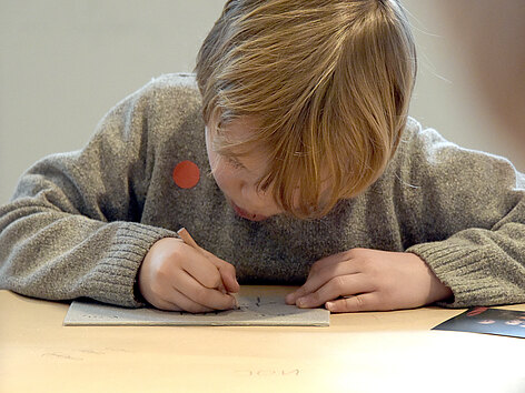 Ein kleiner Junge sitzt hinter einem Tisch und beugt sich über ein Blatt Papier, auf das er mit einem Stift etwas malt.