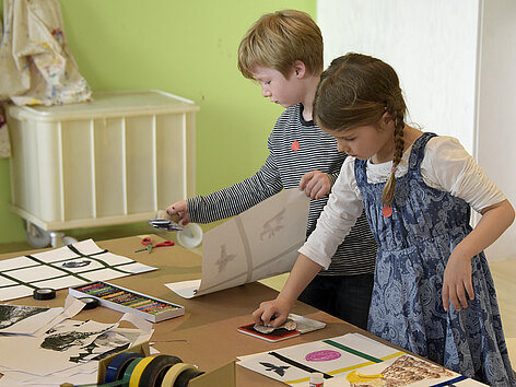 Workshop: Ein Mädchen und ein Junge bedrucken an einem Tisch stehend Papierbögen mit bunten Mustern.