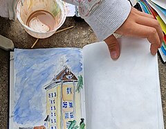 Eine Kinderhand, die in einem Skizzenblock blättert. Man sieht eine Zeichnung vom alten Hochhaus an der Klemensbrücke in Kaiserswerth