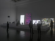 Im Innenraum der Kunsthalle stehen mehrere Personen vor einem länglichen Podest, auf dem verschiedene Projektoren stehen, die Bilder auf die Wand projiziern.