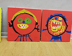Zwei Selbstportraits von Kindern aus einer Kita. Auf zwei Holzplatten, ca. 60x 60 cm groß, sie sind an eine Wand gelehnt. Die Platten sind rot grundiert und darauf sind die Portraits gemalt. Sehr bunt, mit gelb, grün, blau und weiß.