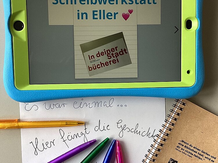 Tablet mit Text "Schreibwerkstatt in Eller", davor bunte Kugelschreiber, Blatt Papier mit Handschrift "Hier fängt die Geschichte an" und kleines Notizbuch.