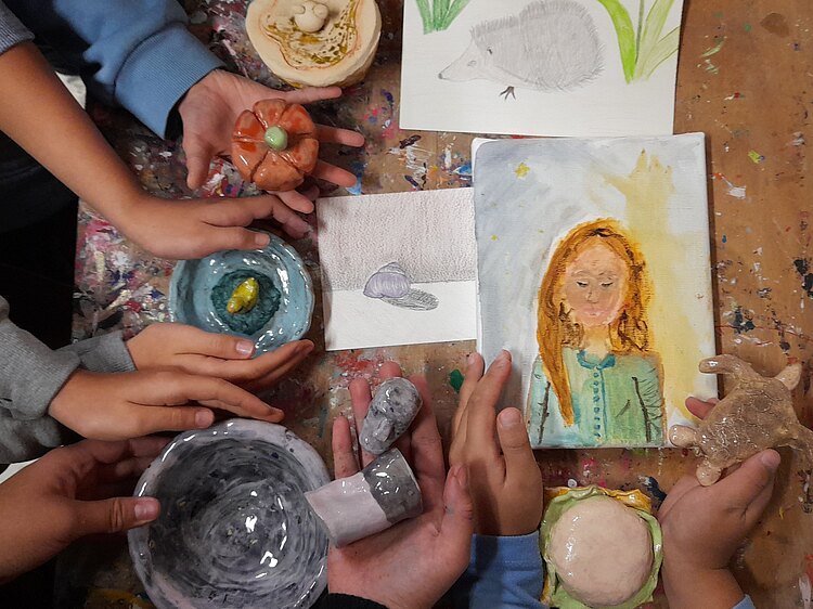Auf dem Foto sieht man Kinderhände und Hände eines Erwachsenen, die gebrannte und glasierte kleine Tonschalen in der Hand halten. Auf dem Tisch liegen verschiedene bunt gemalte Zeichungen vvon einem Gesicht oder einem Igel.