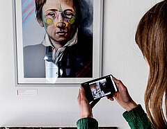 Person mit Smartphone steht vor einem Porträt Heinrich Heines