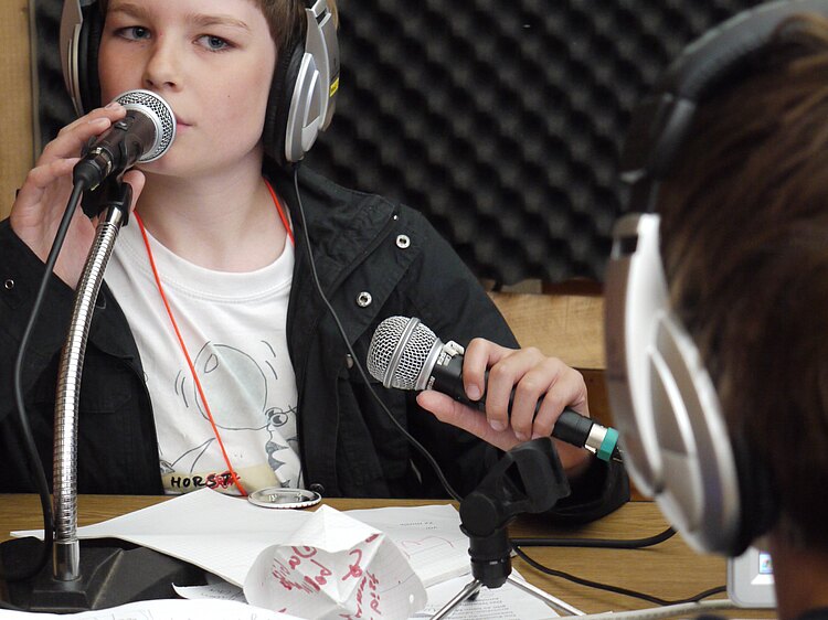 Junge mit Kopfhörern am Mikrofon in Technikkabine mit anderem Kind als Gegenüber