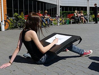 Junge Frau sitzt auf dem Boden in einer Fußgängerzone mit einer großen Unterlage auf dem Schoß und zeichnet