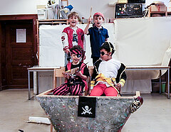 Vier Jungs als Piraten verkleidet hinter ihrem Schiff, das ein umgedrehter Dreieckstisch ist. Dekoriert ist das Schiff mit einer Piratenflagge Piratenflagge daran