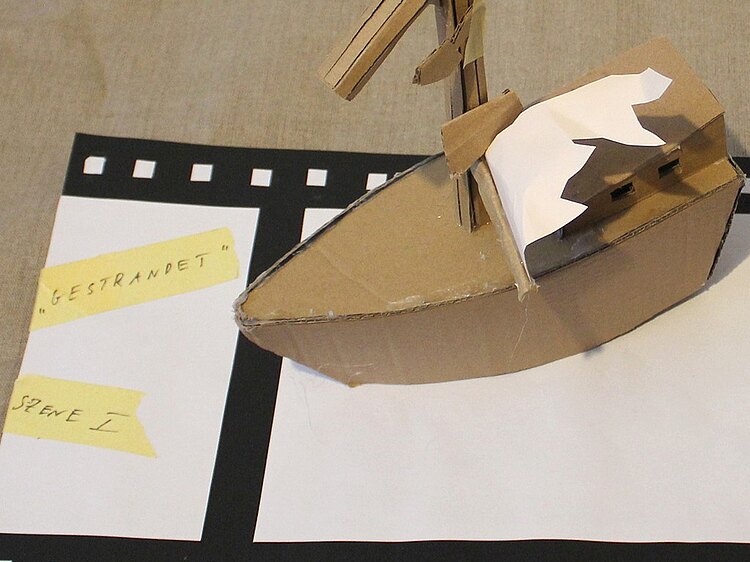 Ein Schiff, das aus Pappe gestaltet wurde, steht auf einer ebenfalls aus Pappe gestalteter farblich angemalter Filmrolle